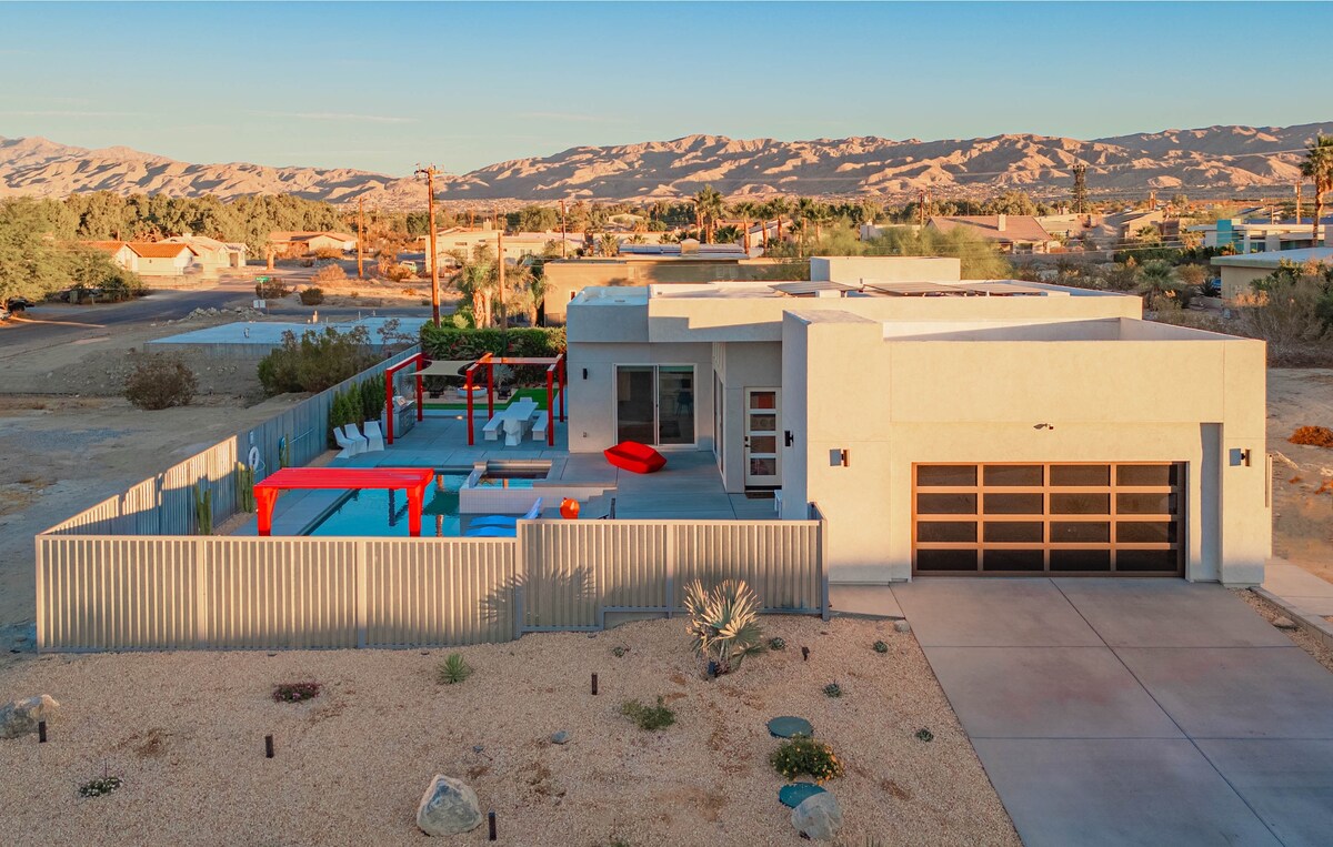 An aerial view of StarHaven showcases its modern architecture set against a serene desert backdrop. The patio features a private pool, lounge seating, and vibrant red accents. A spacious driveway is visible leading to the garage, with surrounding desert landscaping and hills in the distance.
