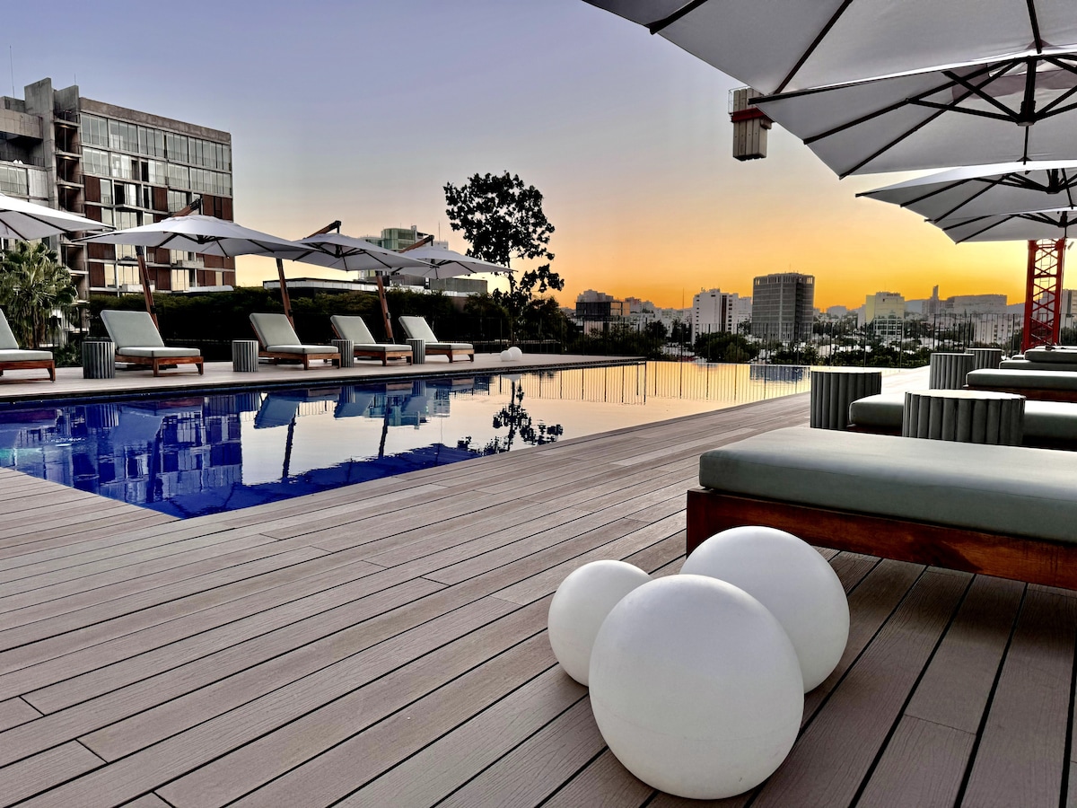 A serene pool area features a sleek, reflective surface surrounded by comfortable lounge chairs and large umbrellas. The evening sky showcases warm hues as the sun sets, while minimalist white sculptures add a modern touch to the wooden deck.