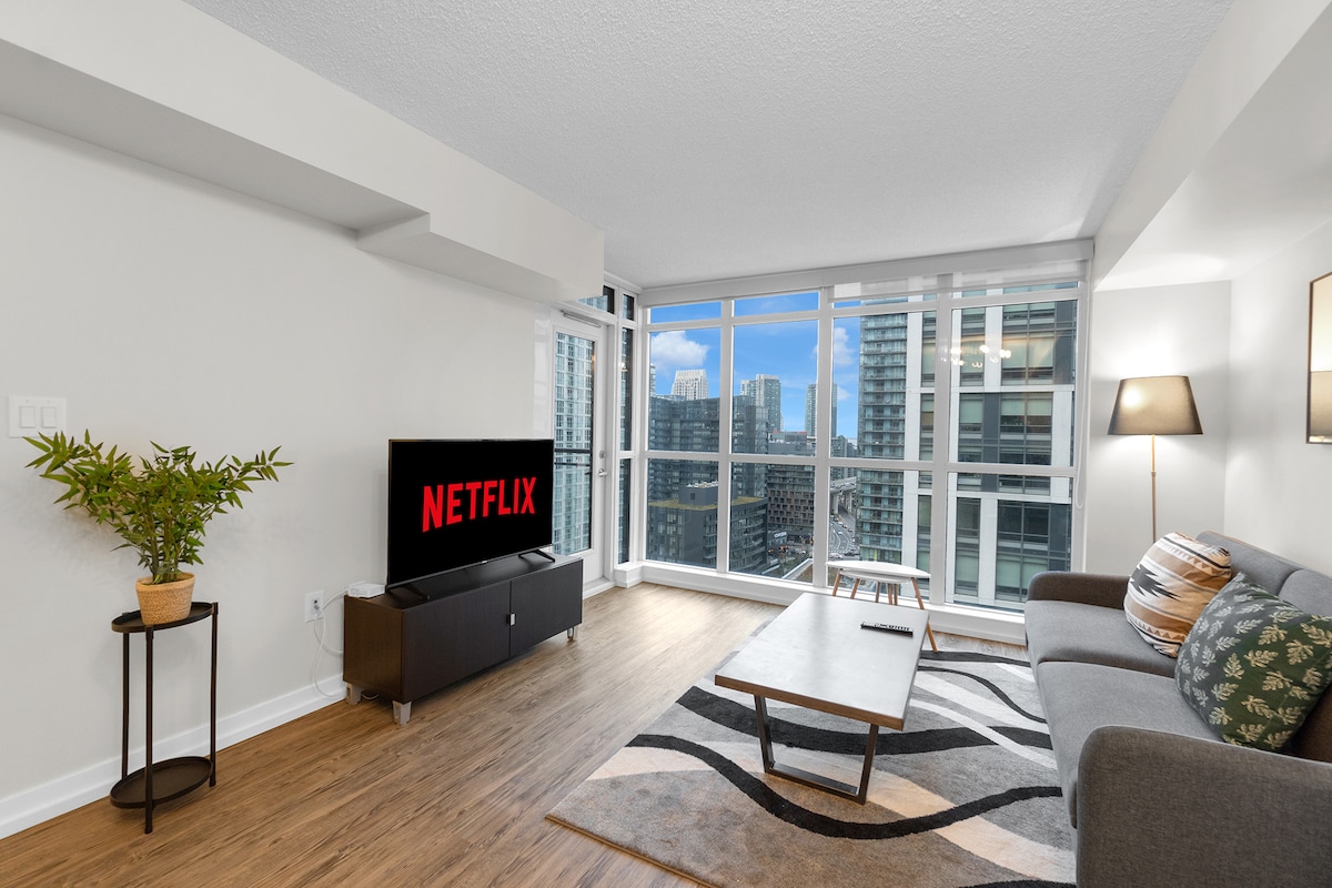 A modern living area is shown with a sleek sofa facing a flat-screen television displaying the Netflix logo. Large windows allow natural light to illuminate the space, revealing a gentle view of the surrounding cityscape. A stylish coffee table complements the decor, with a small potted plant adding a touch of greenery.