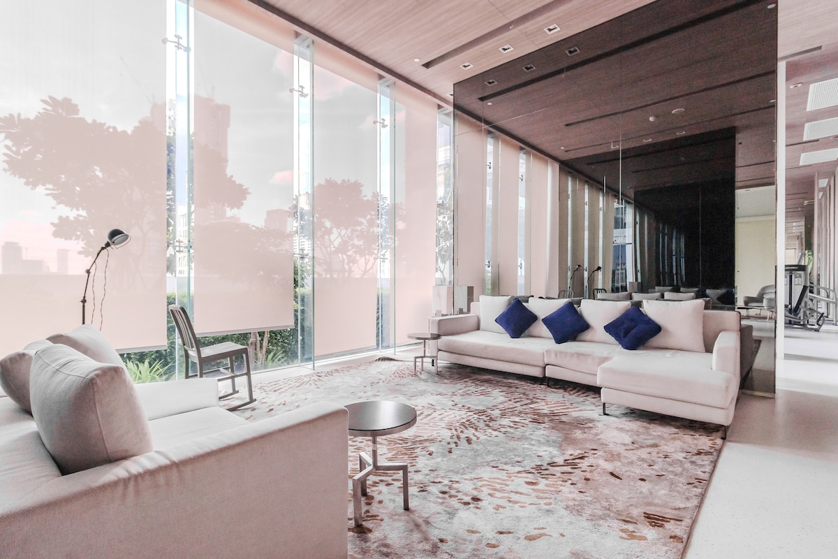 A modern lounge area features large glass windows allowing natural light to fill the space. Comfortable seating options include a light-colored sofa and armchairs, complemented by decorative blue cushions. A round table is positioned in the center, with a textured area rug adding warmth to the room.