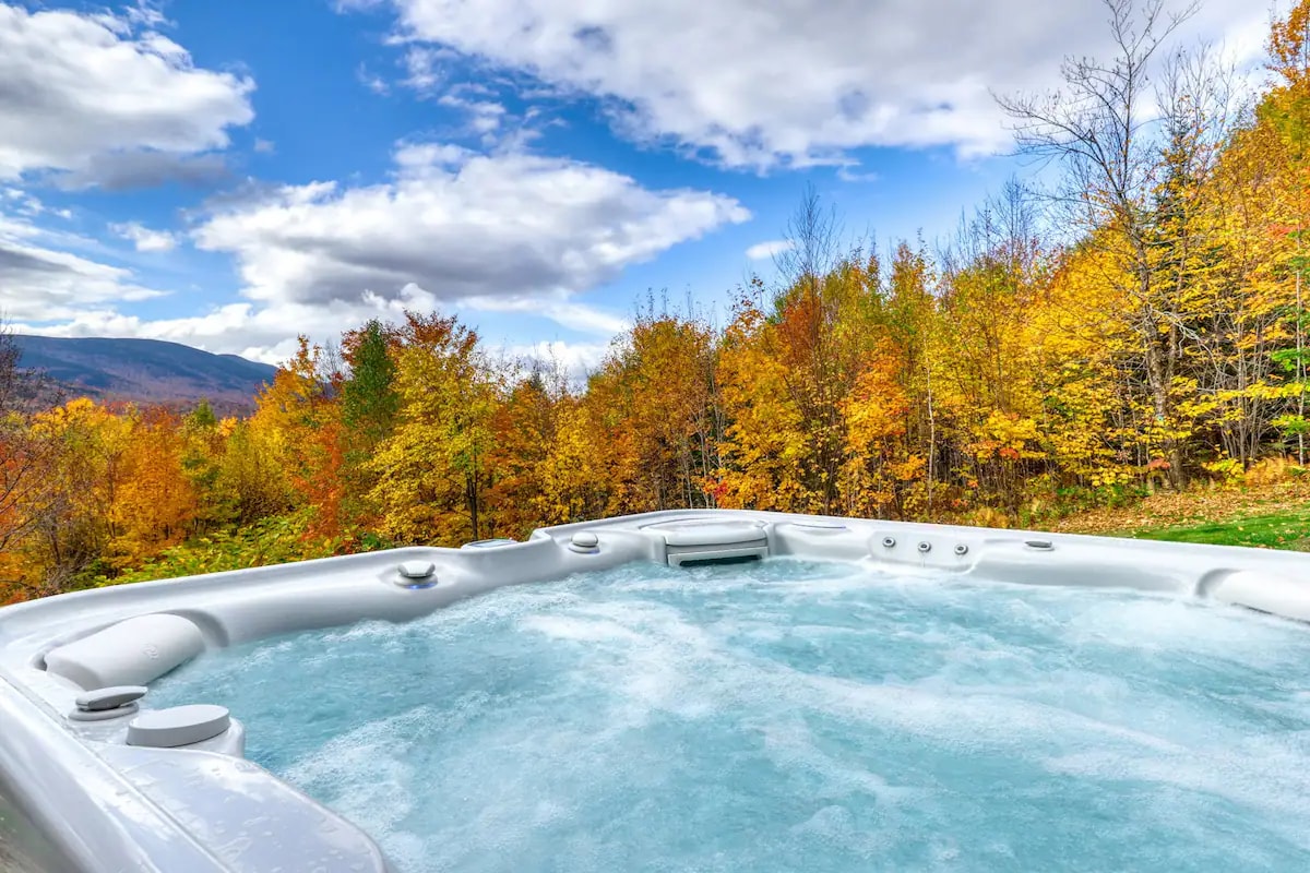 A hot tub filled with bubbling water is positioned outdoors, surrounded by vibrant autumn foliage. Warm colors of orange, yellow, and red enhance the natural setting, contrasting with a clear blue sky scattered with clouds.