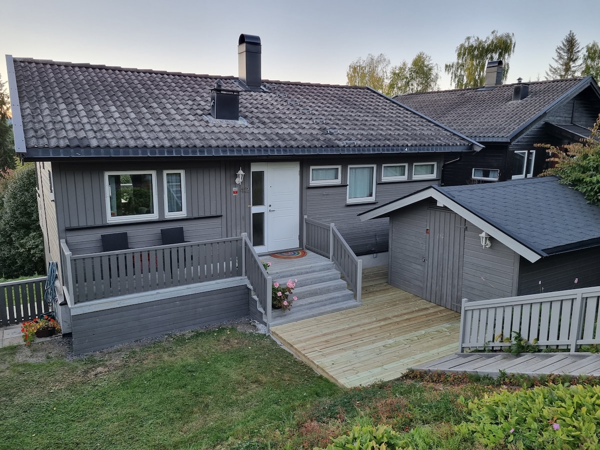 A charming exterior of a house is presented, showcasing a sloped roof with tiles. A wooden deck leads to the entrance, flanked by two seating areas. Surrounding greenery is visible, along with a storage shed subtly incorporated into the landscape.