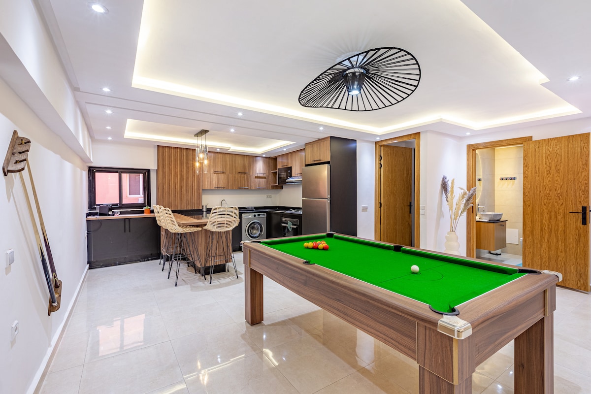 A spacious living area features a billiard table at its center, surrounded by modern decor. The kitchen area is visible in the background, equipped with appliances and bar seating. Soft lighting accents the ceiling, providing a bright and welcoming environment.