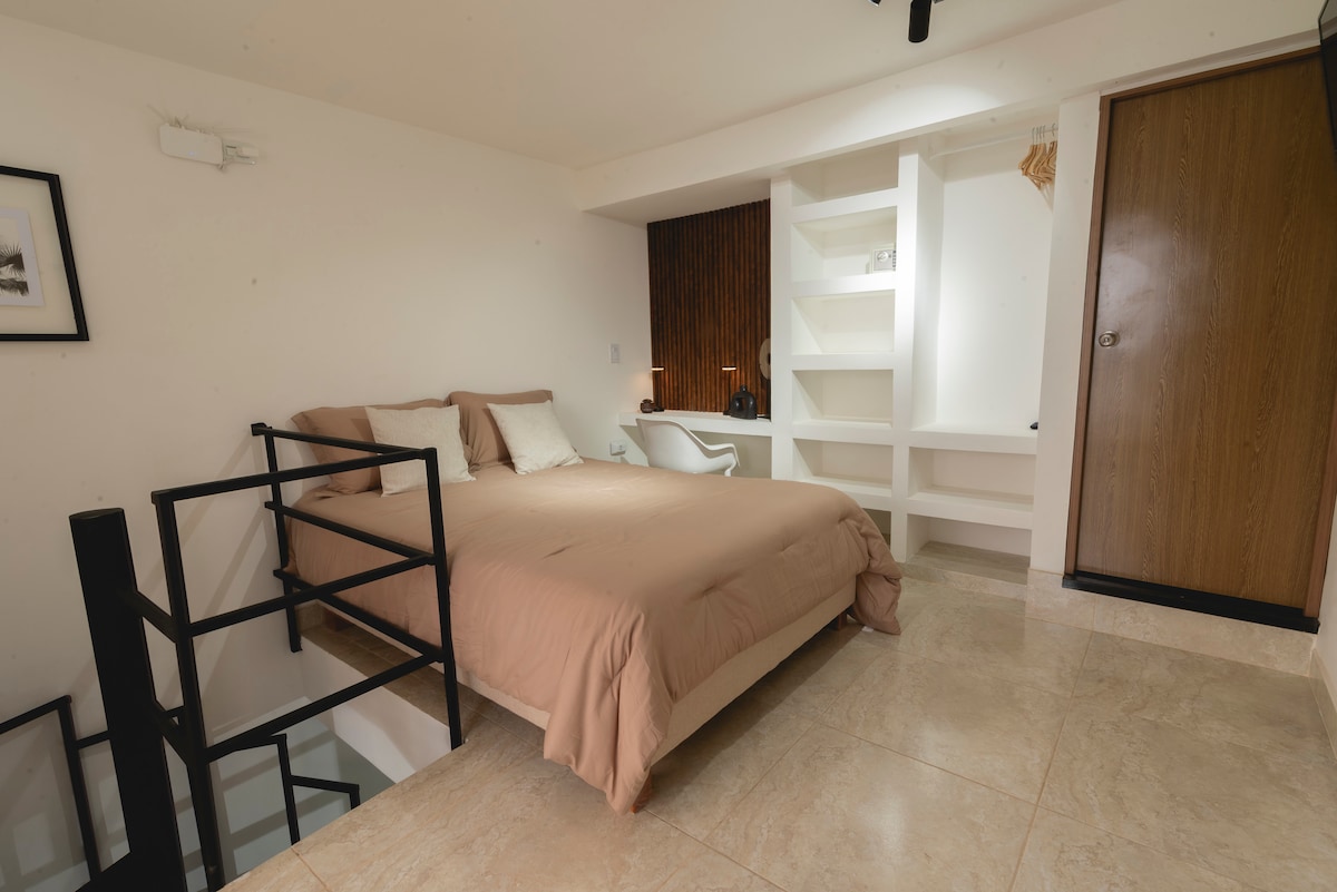 The cozy bedroom features a spacious double bed with neutral bedding. Soft lighting enhances the calm ambiance. A compact workspace is available nearby, complemented by minimalist decor. The room is accessed via a modern spiral staircase, which adds a stylish touch to the space.