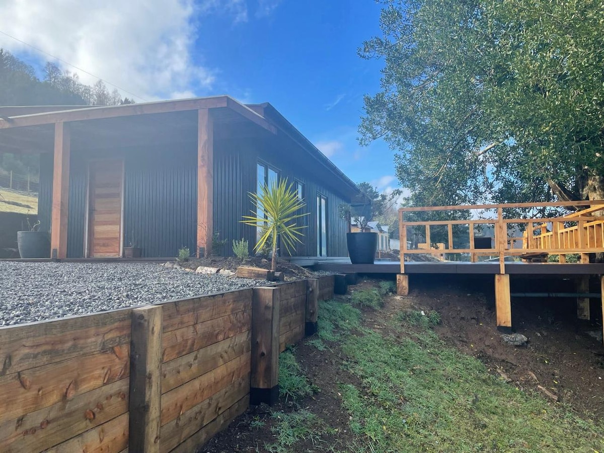 A modern cabin is set against a blue sky, featuring a combination of natural wood and dark exterior materials. A wooden deck extends from the front, surrounded by landscaped areas with potted plants and gravel pathways leading up to the entrance.