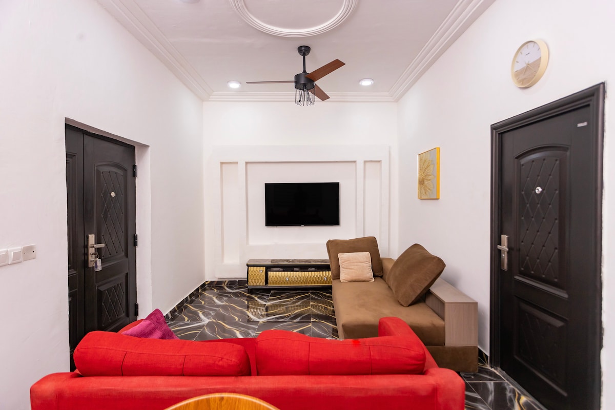 A modern living room features a sleek wall-mounted TV above a stylish media console. Comfortable seating includes a brown sectional sofa and a red couch. A ceiling fan and overhead lights provide illumination. The glossy dark floor complements the white walls and decorative accents.