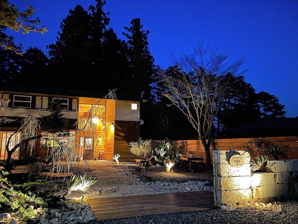 The exterior of the guesthouse features a warm light illuminating the entrance against a deep blue night sky. A wooden pathway leads to the entrance, bordered by plants and stones, creating a serene and inviting atmosphere amidst surrounding trees.