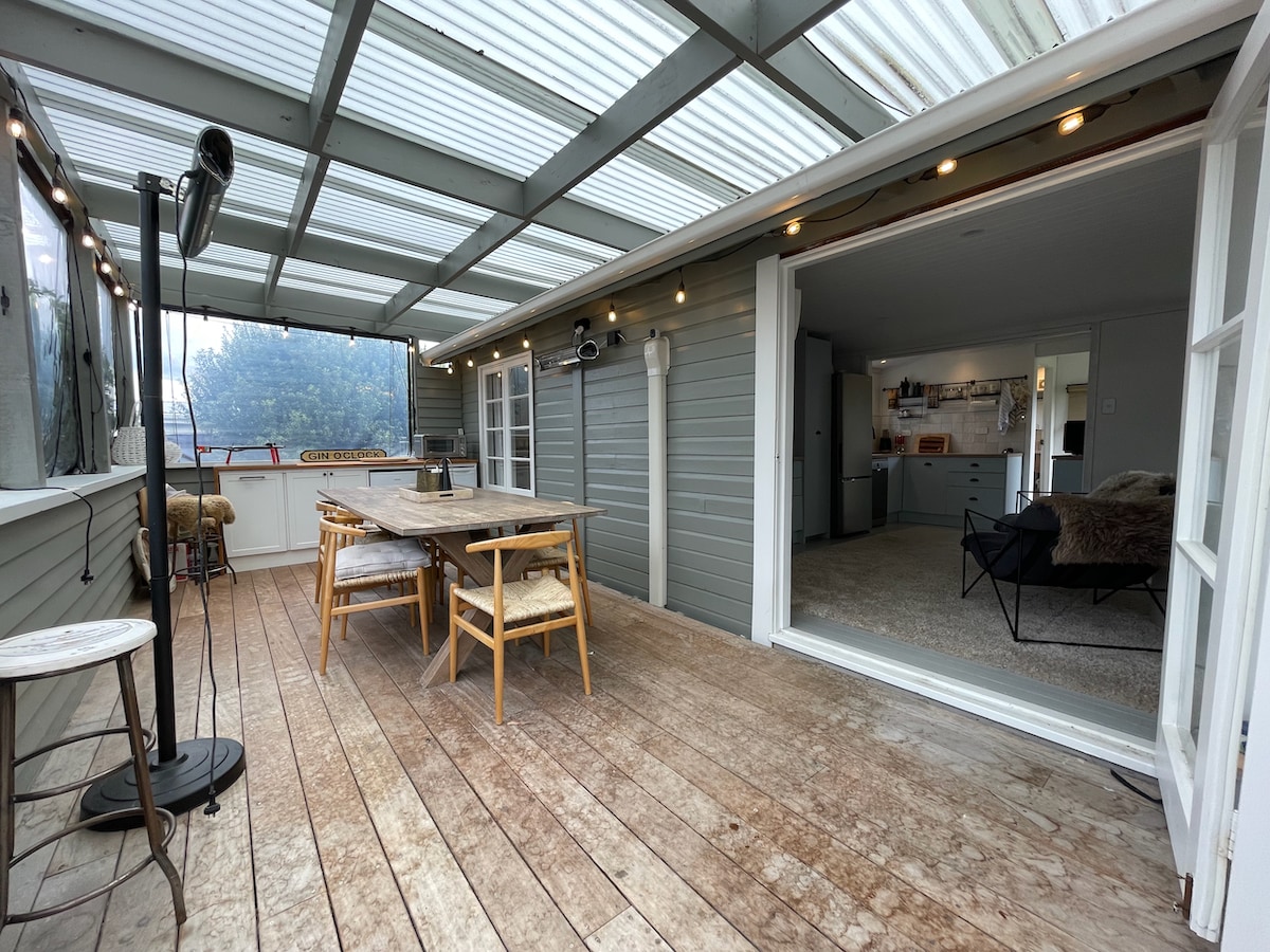 A spacious deck area features a wooden dining table surrounded by four chairs, illuminated by soft lighting. Large glass doors open to an inviting indoor space, allowing natural light to flow in. The wooden floorboards add a rustic feel to the environment.