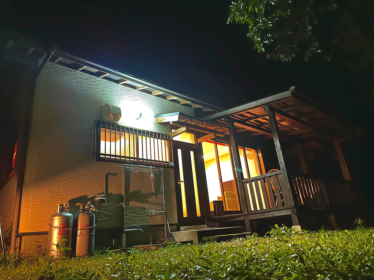 The exterior of the villa is illuminated at night, highlighting the wooden porch and entrance. Feature details include outdoor lighting, gas tanks, and a well-maintained lawn. Large windows showcase a warm glow from inside, suggesting a cozy atmosphere.