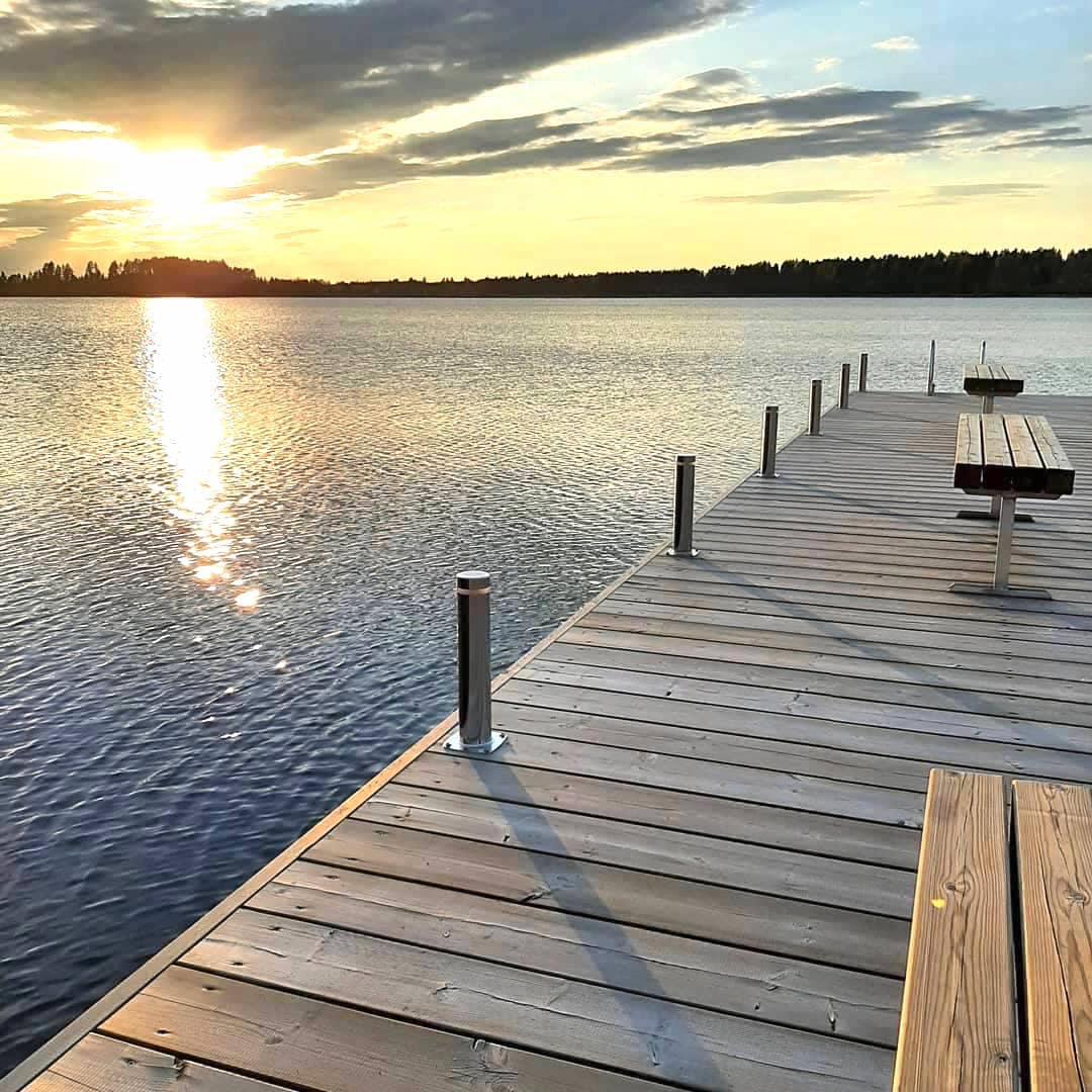 A wooden dock extends over calm waters, reflecting the warm hues of a sunset. The shoreline is lined with trees in the background, and a picnic table is positioned at the edge of the dock, inviting guests to enjoy the serene views.