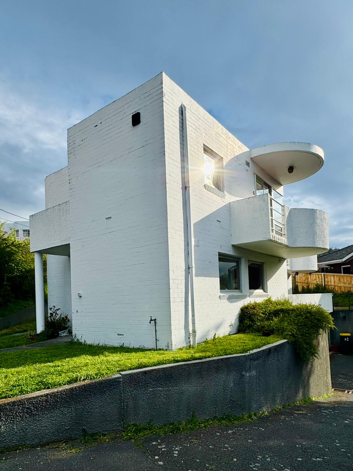A unique three-level white townhouse is displayed, showcasing a modern architectural design. Large windows provide ample natural light, while a curved balcony adds character to the structure. Lush green grass surrounds the base, contributing to the serene environment.