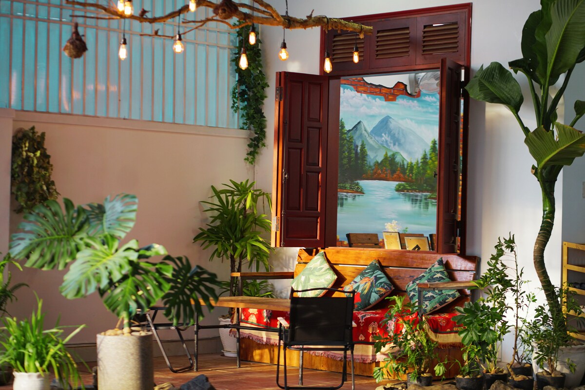 A cozy sitting area features wooden furniture adorned with colorful cushions. Sunlight filters through open doors, illuminating a mural depicting mountains and water. Lush green plants and decorative lighting enhance the space, creating a calming environment.