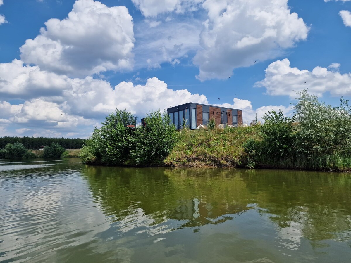 A modern building is situated on a grassy hilltop beside calm water. Lush greenery frames the structure, while fluffy clouds float in the blue sky above. The scene reflects tranquility, highlighting the natural surroundings that create a serene atmosphere.