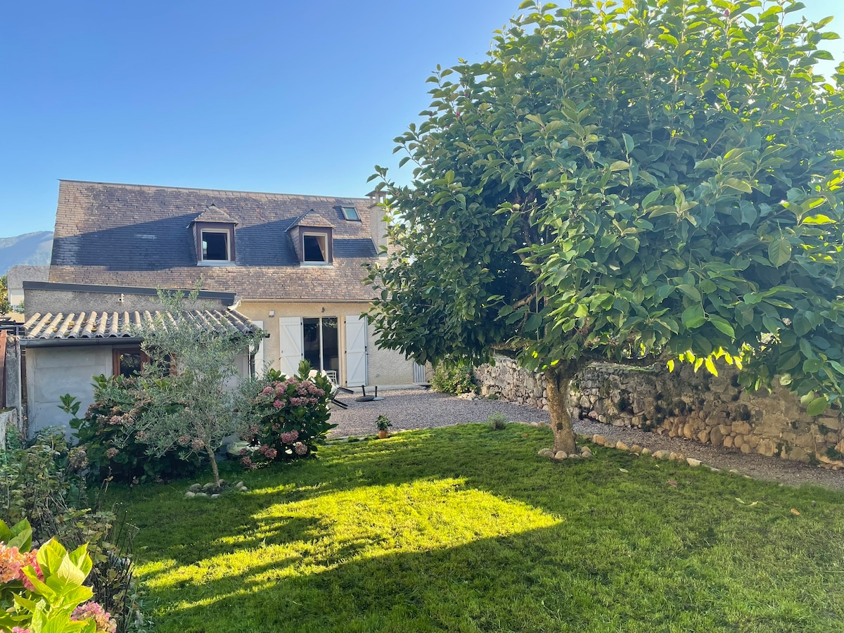 A spacious exterior view showcases a well-maintained garden featuring lush green grass and blooming shrubs. A large tree provides shade, while the house, with its distinctive rooflines and multiple windows, opens invitingly to the outdoors. A stone wall borders the property.
