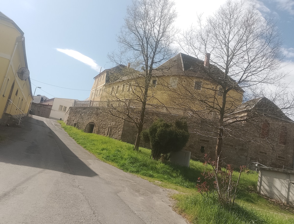 A historic building is positioned at the end of a gently sloping road, surrounded by grassy fields and trees. The structure features a combination of stone and yellow facade, with windows framed by dark shutters. A clear sky is visible above, enhancing the serene setting.