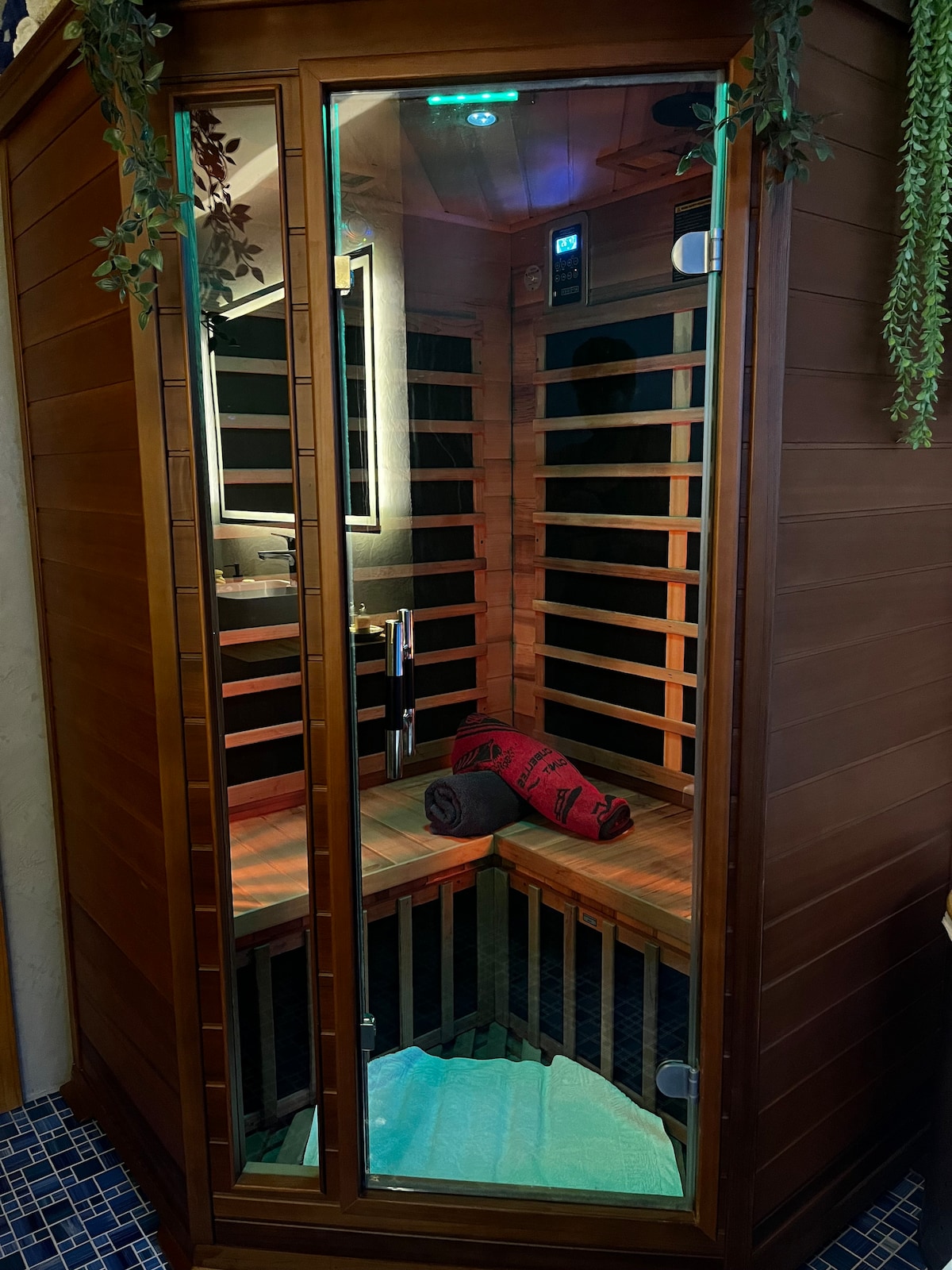 An infrared sauna is presented, featuring a glass door that allows visibility into the wooden interior. The sauna is equipped with wooden benches arranged in a corner configuration, complemented by a towel resting on one of the benches. Soft lighting enhances the serene environment.