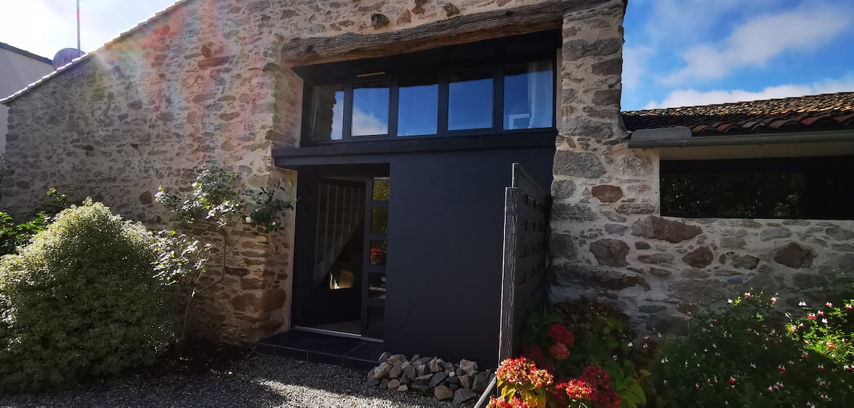 The image features the entrance of a renovated stone building, showcasing a modern black door framed by large windows. Surrounding greenery and colorful flowers add a natural touch to the space, while gravel pathways lead to the entrance, inviting guests into the retreat.