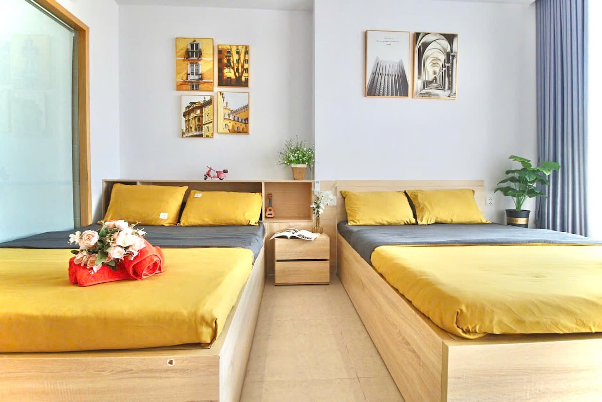 The bedroom features two spacious beds dressed in yellow linens, complemented by grey accents. A small bedside table holds decorative items and a bouquet of flowers. Artwork adorns the walls, and natural light filters in through a window, enhancing the calming atmosphere.