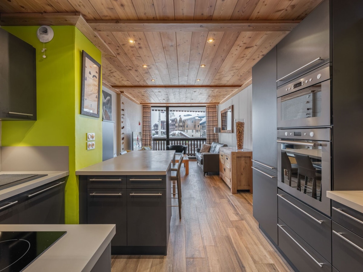 The kitchen features modern cabinetry and appliances, including a built-in oven and induction cooktop. A spacious island offers additional countertop space, while large windows provide natural light and views of the surroundings. Dining space is visible with a table that seats six.