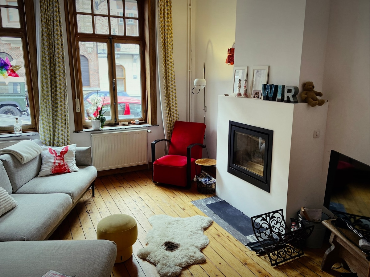 A light-filled living area features a comfortable gray sofa and a red armchair positioned near a modern fireplace. Natural wooden flooring adds warmth, while curtains frame the large windows. A decorative table and a cozy area rug complete the space, creating an inviting atmosphere.