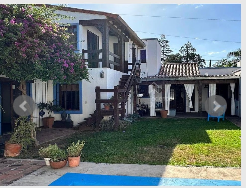 A rustic courtyard is framed by vibrant green grass and flower pots. An exterior staircase leads to a second-floor balcony, while white curtains provide privacy for an adjoining space. The architecture reflects a blend of traditional and inviting design elements.