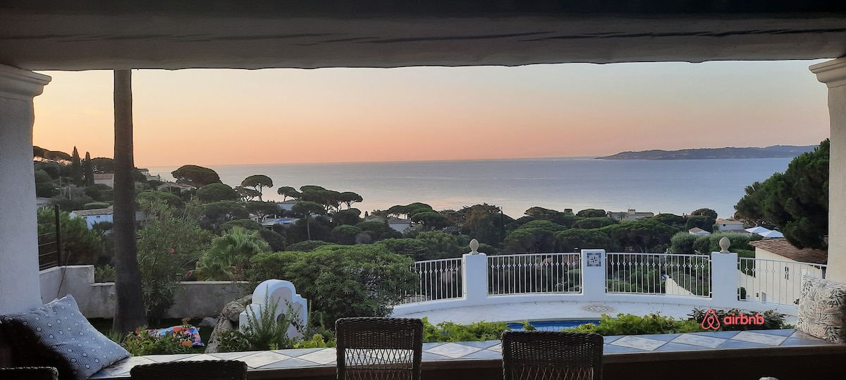 A panoramic view of the coastline is captured at sunset, showcasing serene water hues and lush greenery. The foreground includes a terrace with comfortable seating, allowing guests to enjoy the tranquil environment and beautiful scenery.