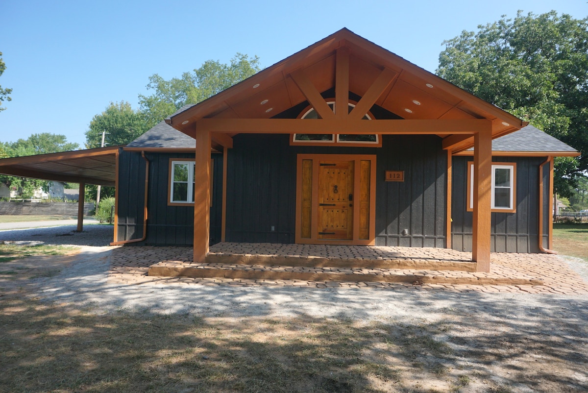 The exterior of a uniquely designed house is showcased, featuring a large wooden door and a gabled roof. The facade combines dark siding with warm wooden accents. A spacious covered area is seen at the side, while a gravel path leads to the entrance, surrounded by a grassy area.