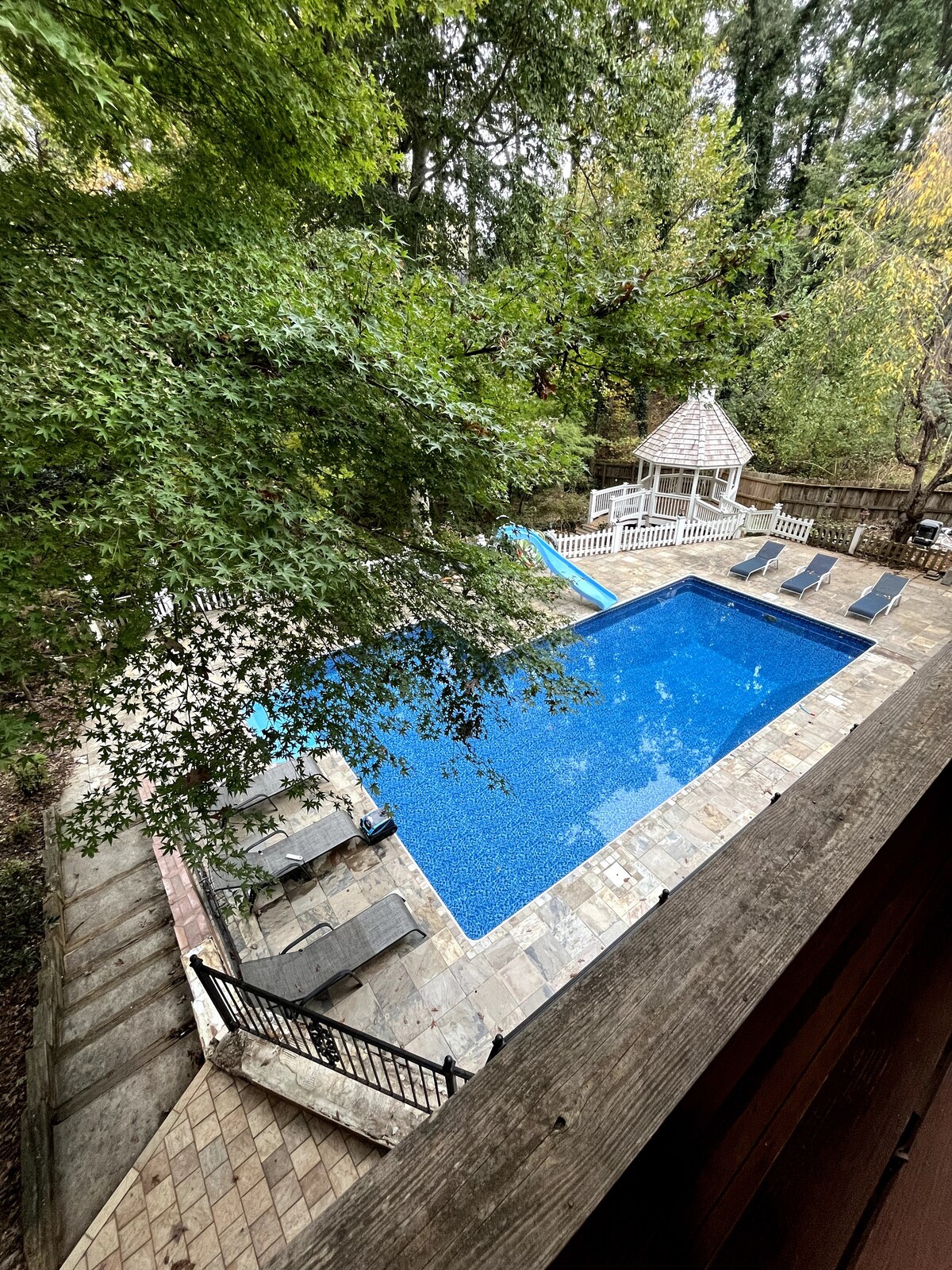 An expansive pool area is visible, featuring crystal-clear water surrounded by a stone patio. Several lounge chairs are positioned around the pool, and a covered gazebo is located at one end. Lush trees provide natural shade and privacy, enhancing the outdoor experience.