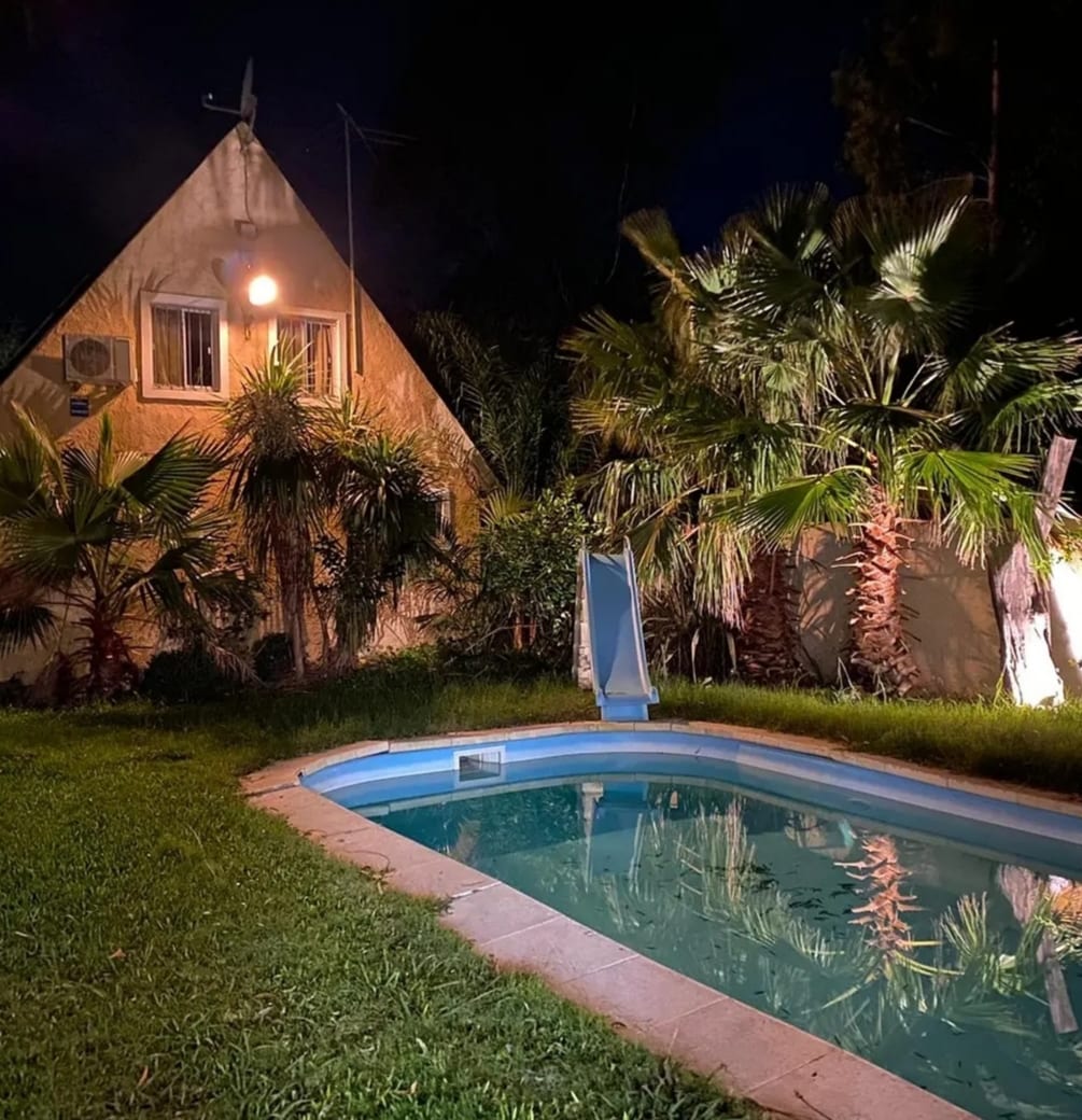 A triangular-shaped house is illuminated at night, surrounded by tropical vegetation. A small, unheated pool features a slide, and lush green grass frames the scene, creating a serene outdoor environment.