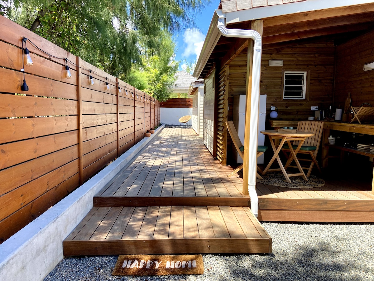 A wooden deck path leads to the entrance of the lodge, framed by a privacy fence. To one side, an outdoor kitchen is visible, featuring essential appliances and a small dining table. Decorative lights create a welcoming ambiance, while a sun lounger rests nearby.