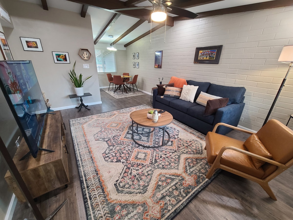 A comfortable living area features a navy sofa adorned with decorative pillows, accompanied by a mid-century armchair. A circular wooden coffee table sits at the center, while a colorful area rug adds warmth. Natural light enters through a nearby window, highlighting the space's earthy tones.