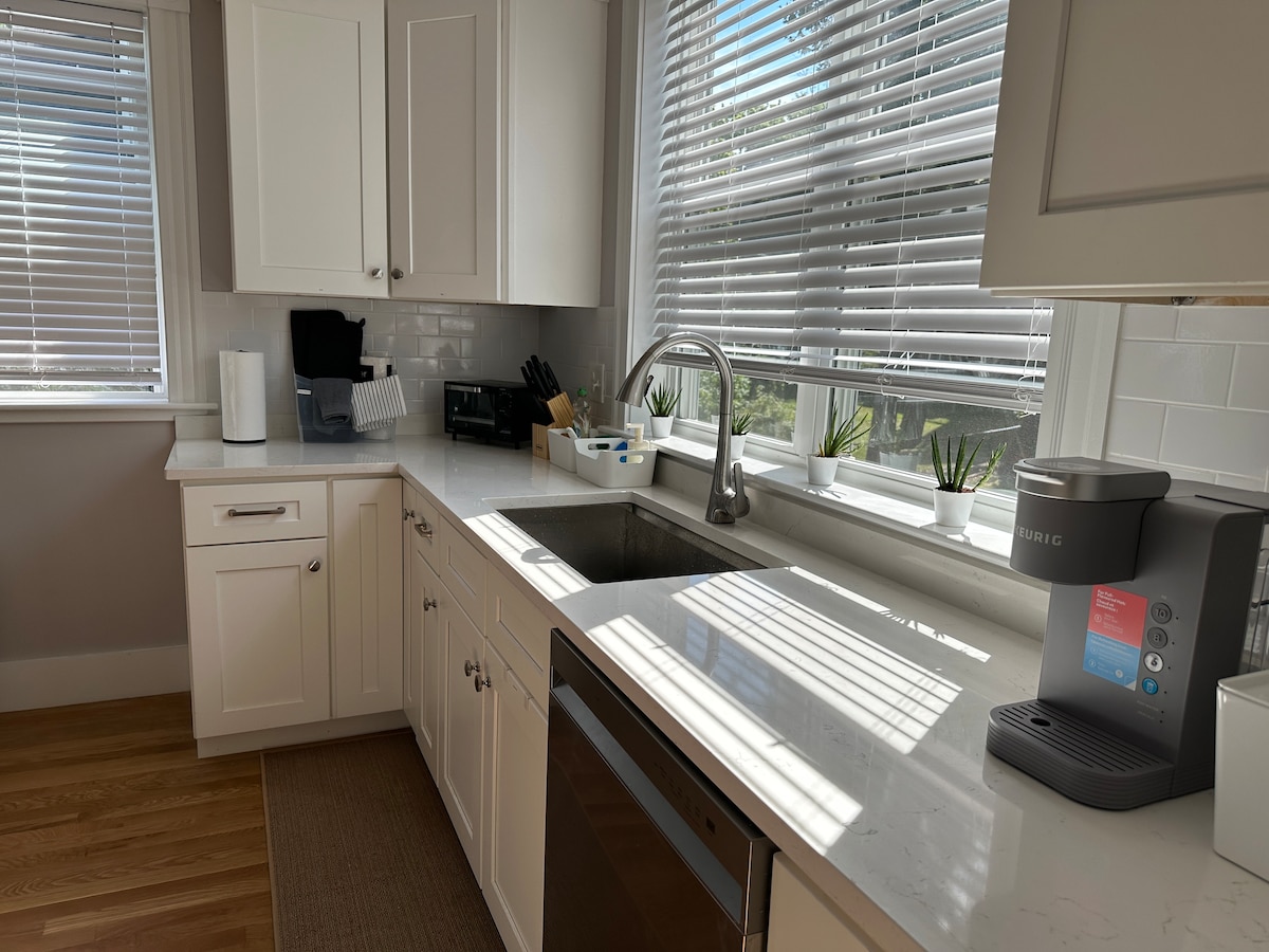 The modern kitchen features white cabinetry and a spacious countertop, illuminated by natural light through the window. A stainless steel sink is positioned next to a sleek coffee maker, with small potted plants adding a touch of greenery.