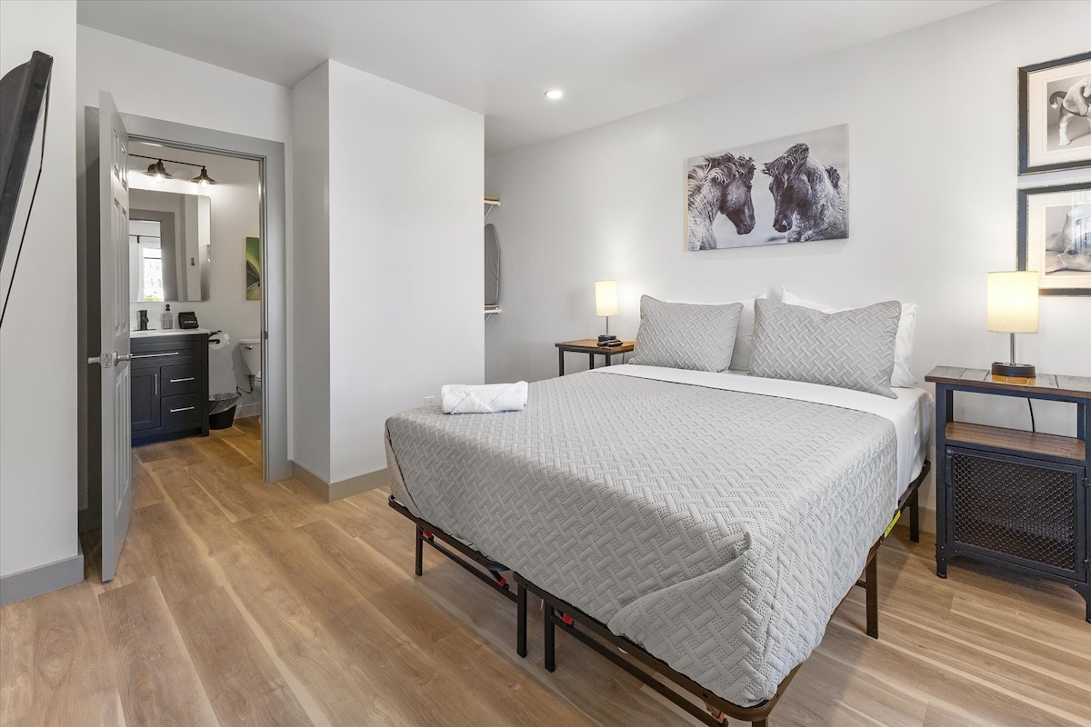 A spacious bedroom features a queen bed with a textured gray blanket and four pillows. Two lamps provide soft lighting on either side, while a closet is visible in the background. The entry to a modern bathroom can be seen in the distance.