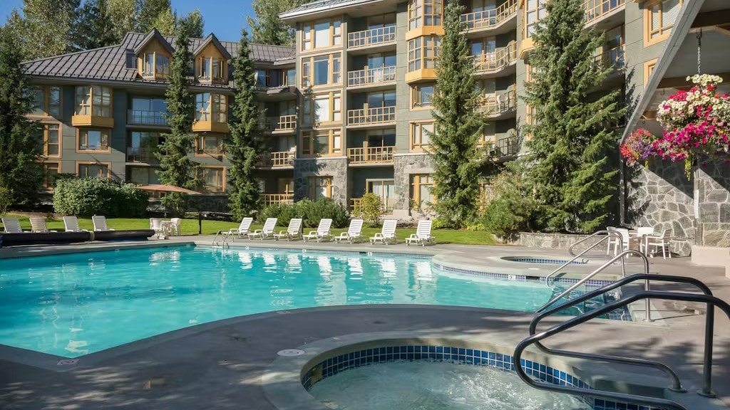 A spacious outdoor pool area is surrounded by tall trees and well-maintained landscaping. Sun loungers are placed around the pool, while a hot tub is located nearby. The inviting pool is sheltered by the lodge, which features multiple balconies and large windows.