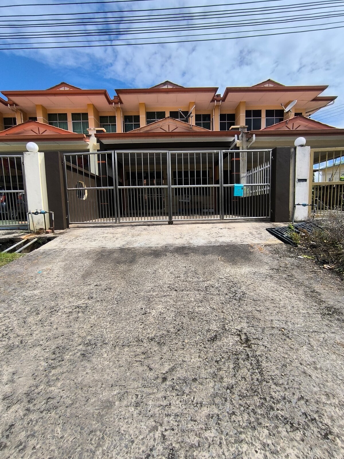 A modern townhouse is prominently featured with a gated entrance. The structure showcases a symmetrical design with a sloped roof and large windows. Sunlight reflects off the gate, highlighting the paved pathway leading up to the entrance.