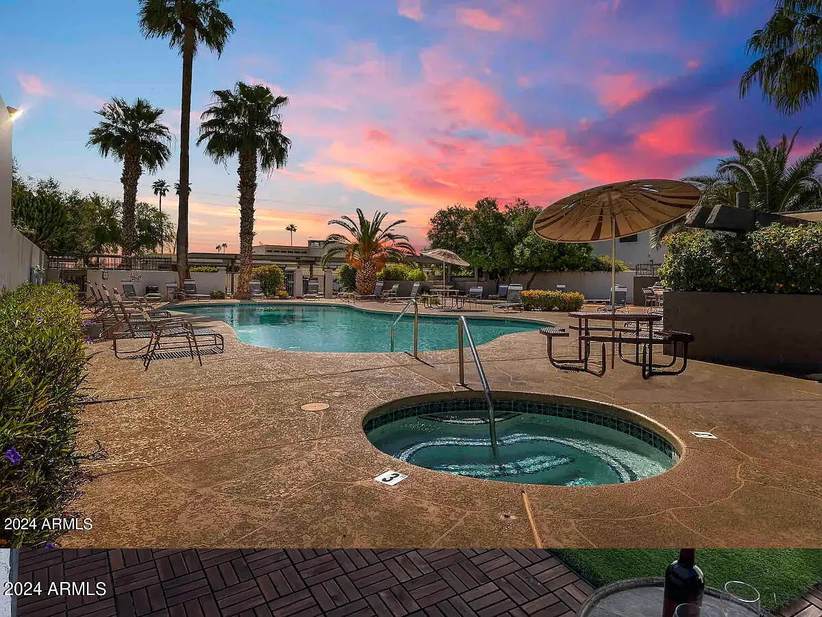 A pool area is surrounded by palm trees and lush landscaping, featuring a spacious swimming pool with a hot tub nearby. Several lounge seats and an umbrella provide shaded seating. The sunset sky showcases vibrant hues of pink and orange, enhancing the outdoor experience.