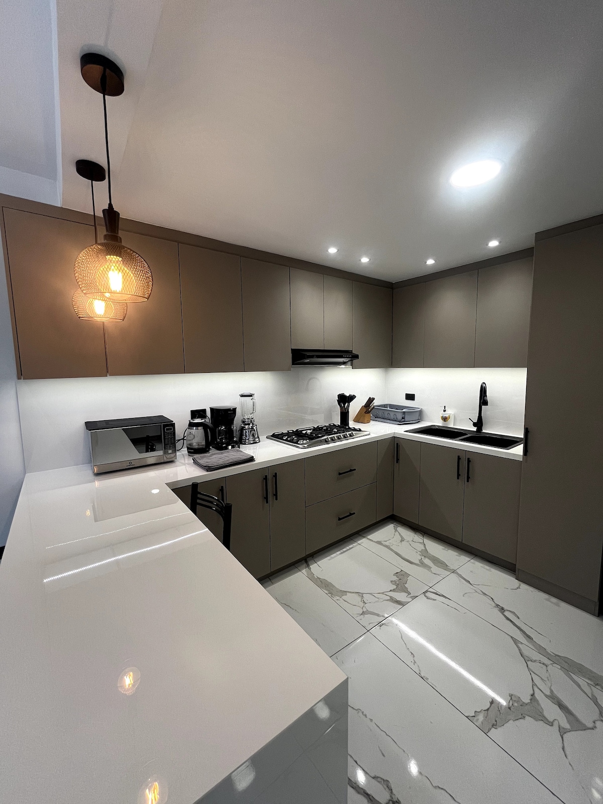 A modern kitchen features sleek cabinetry and a spacious countertop. Stainless steel appliances, including a microwave and coffee maker, are visible. The room is well-lit by recessed lighting and a pendant light, while marble-patterned flooring adds an elegant touch.