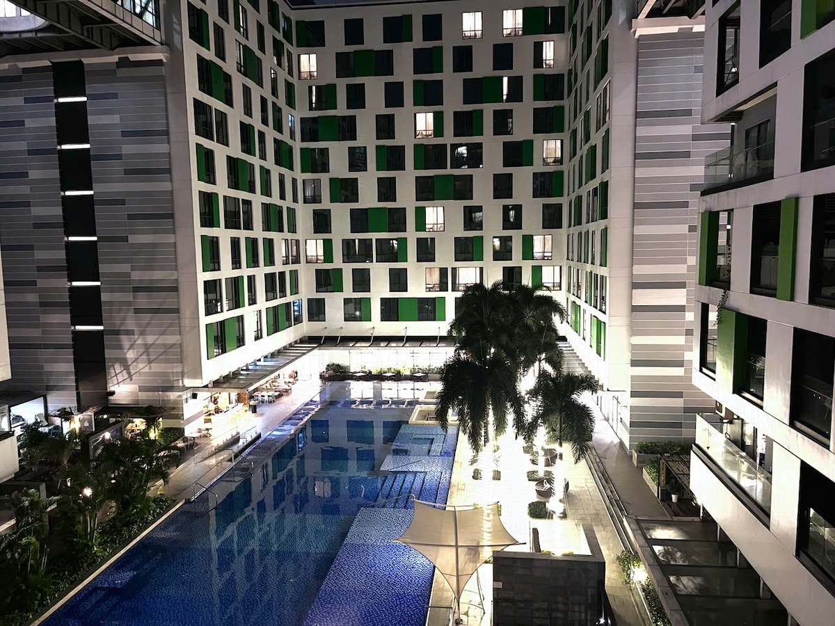 The outdoor swimming pool is surrounded by a stylish building, featuring multiple levels and large windows with green accents. Palm trees and lounge chairs are positioned around the pool area, illuminated at night to create a welcoming ambiance.