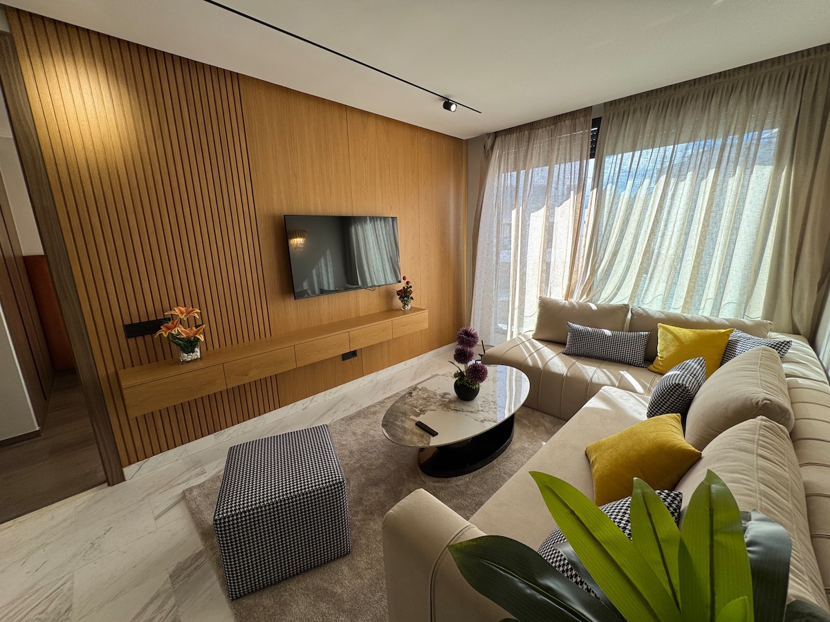 Დახვეწილი მისაღები ოთახი კუთხის კრემისფერი დივნით, ფერადი ბალიშებითა და ხის კედლით კედელზე მიმაგრებული ტელევიზორით. Შუშის ყავის მაგიდა და პუფები კომფორტს ქმნის, შუშის დიდ ფანჯარას კი ლამაზი სიკაშკაშე მოაქვს.