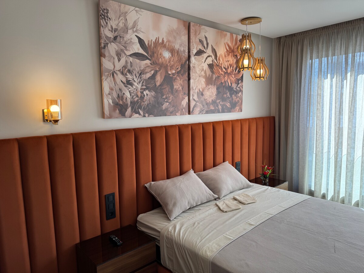 Დახვეწილი ოთახი ორადგილიანი საწოლით, ნარინჯისფერი ხავერდით მოპირკეთებული თავსაფრით და ყვავილების კედლის დეკორით. Კაშკაშა სავალი ნაწილები და შუშის დიდი ფანჯარა თბილი და ნათელი ატმოსფეროა, რომელიც იდეალურია დასვენებისთვის.