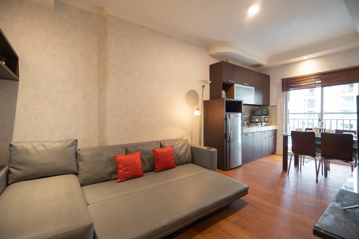 A comfortable living space features a modern sectional sofa adorned with two red cushions. In the background, a sleek kitchen with stainless steel appliances is visible, while a dining table with four chairs is positioned near a window providing natural light.