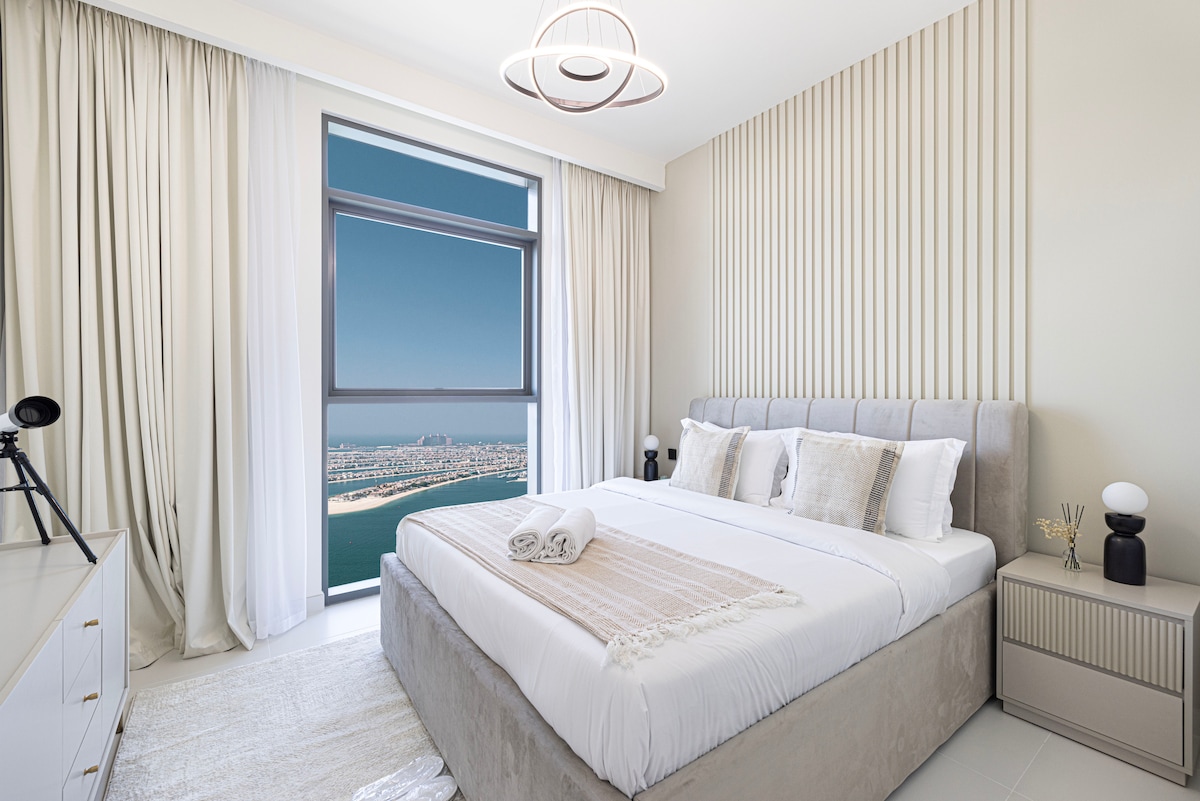 A spacious bedroom is displayed, featuring a plush king-size bed with decorative pillows and a neutral-toned throw blanket. Large windows offer sweeping views of the Dubai skyline and sea, while soft curtains frame the light-filled space. A modern bedside table and lamp are positioned beside the bed.
