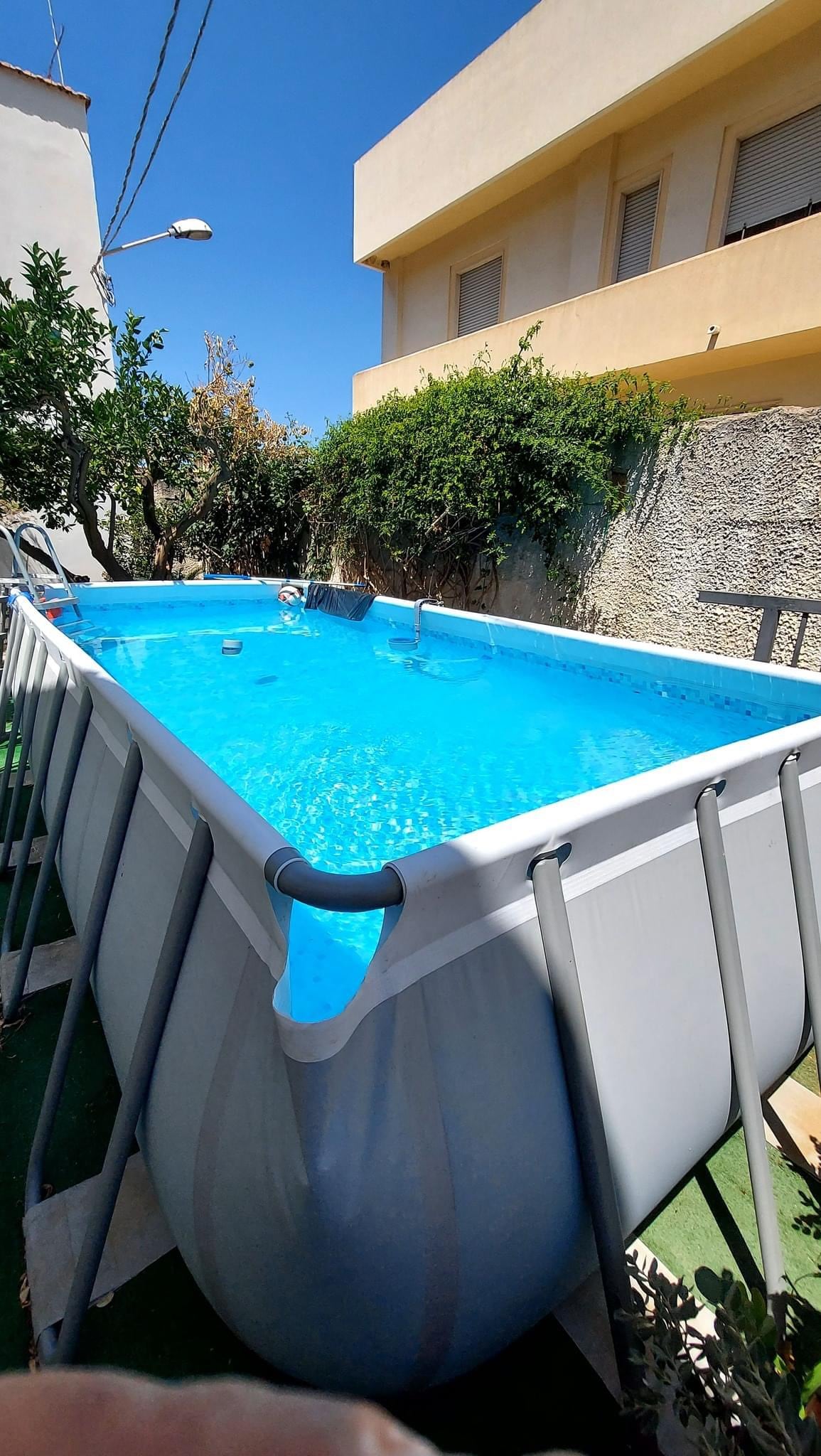 A rectangular above-ground pool is set in a sunlit outdoor area, surrounded by greenery. The clear blue water reflects the sunny sky, while the pool's sturdy frame is clearly visible. Nearby, a ladder provides access to the pool.