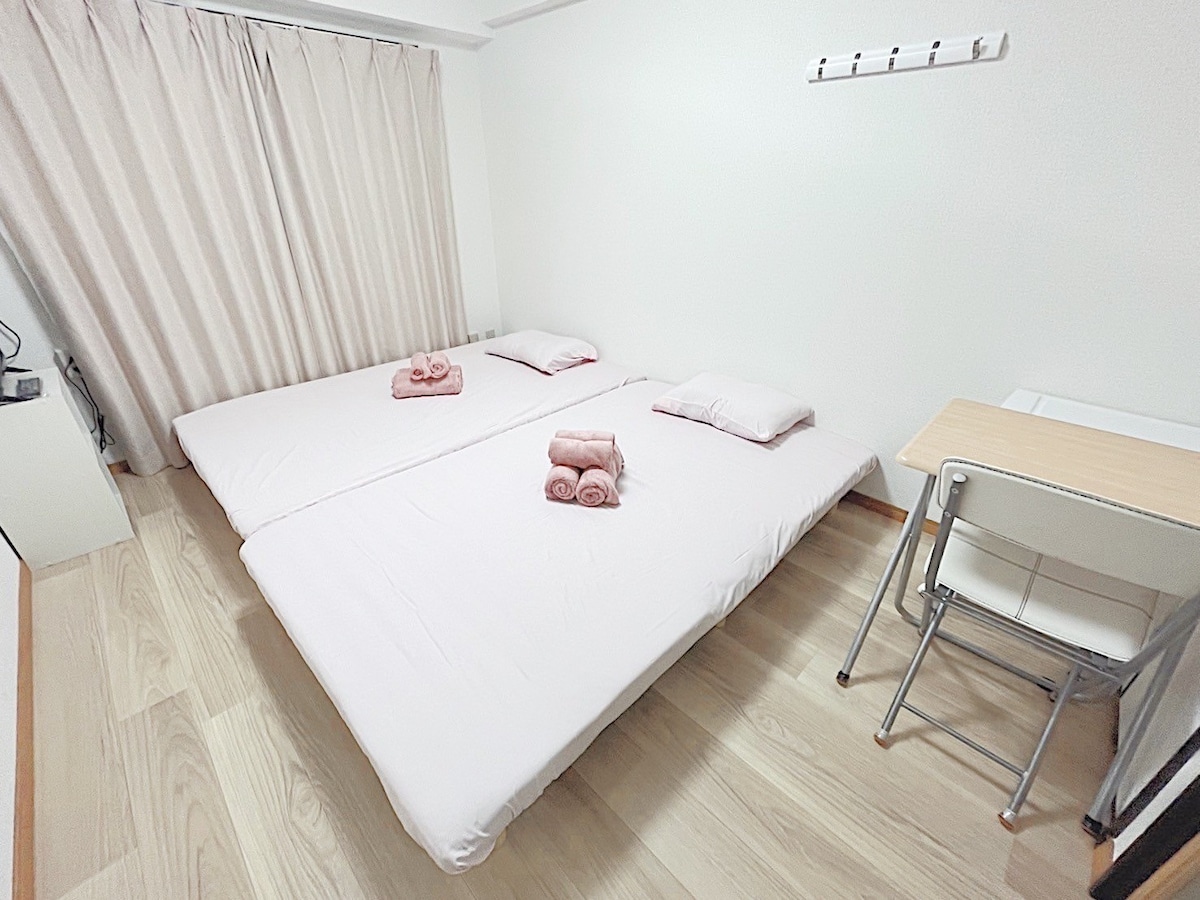 The room features two spacious beds arranged side by side, each topped with rolled towels. A simple desk and chair are positioned against one wall, and light-colored curtains frame a window, contributing to a bright and airy ambiance.