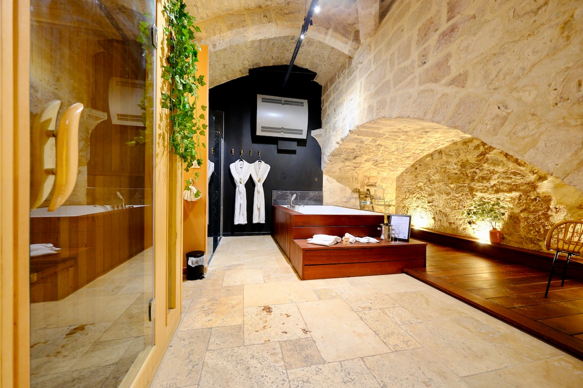 An intimate relaxation area is set within a vaulted stone cellar, featuring a spacious whirlpool bathtub and a sauna. Soft lighting and elegant decor create a calm ambiance. A wooden chair and neatly arranged bathrobes are positioned nearby, enhancing the serene environment.