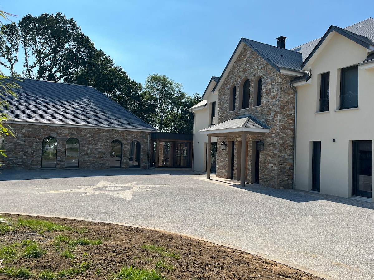 The exterior of a contemporary stone and concrete house is visible under clear skies. The structure features arched windows and a spacious driveway, surrounded by trees. A separate building is positioned nearby, showcasing a cohesive architectural style.