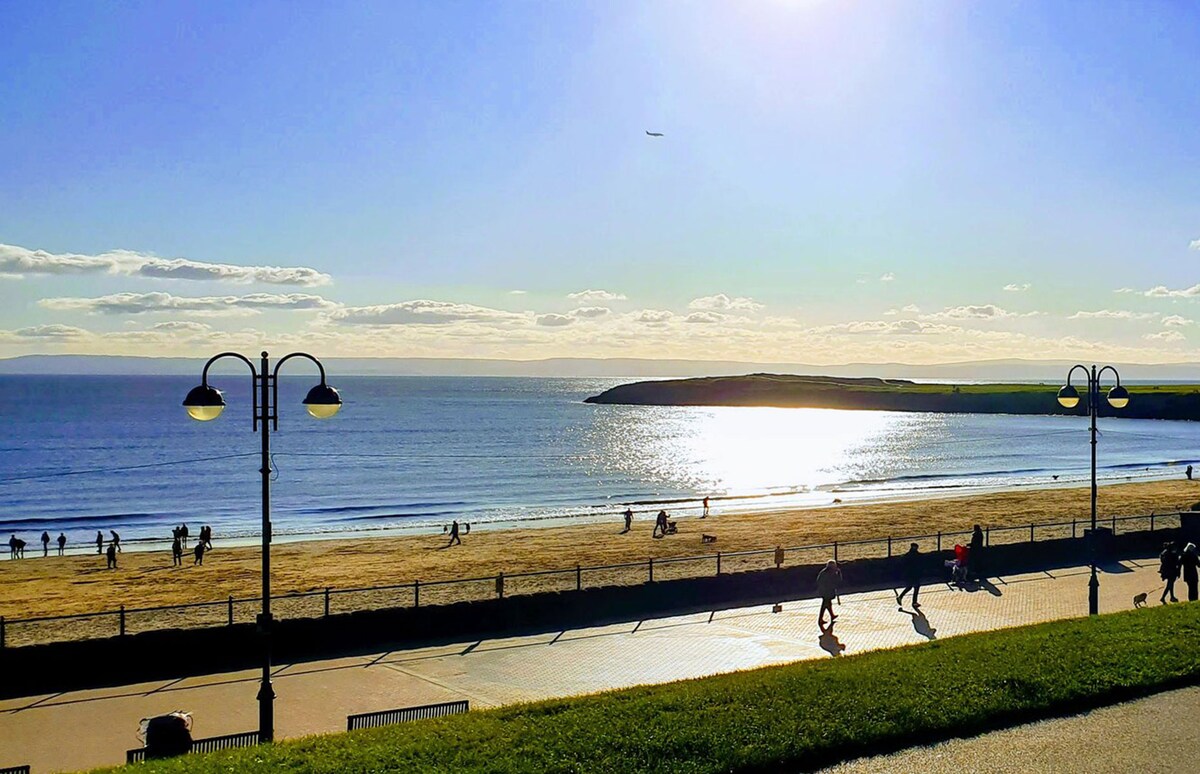 The image captures a scenic beach view, showing a stretch of sandy shore and the sparkling ocean under a clear blue sky. Sunlight reflects off the water, creating a shimmering effect. People can be seen enjoying the beach, contributing to a vibrant atmosphere.