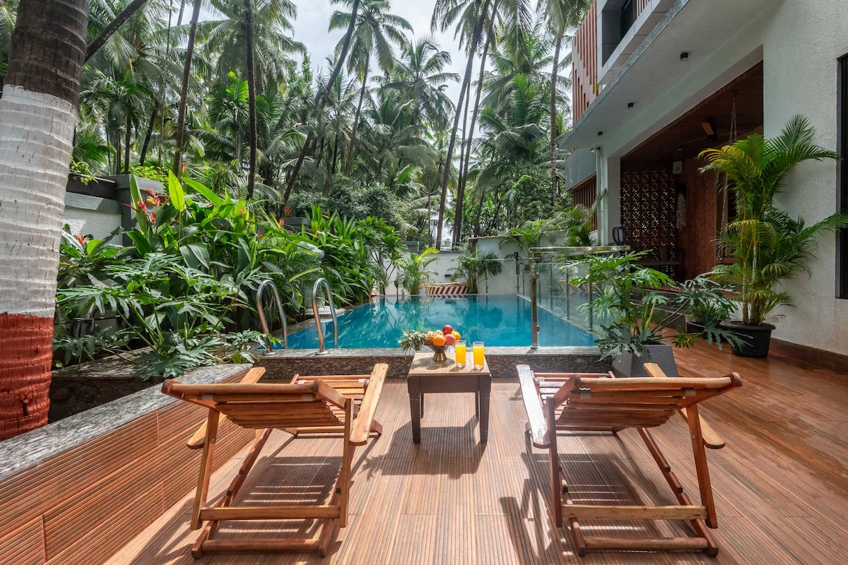 A private outdoor pool is surrounded by lush tropical greenery, creating a serene atmosphere. Two wooden lounge chairs are positioned nearby, alongside a small table with refreshments. Palm trees provide natural shade, enhancing the tranquil setting for relaxation.