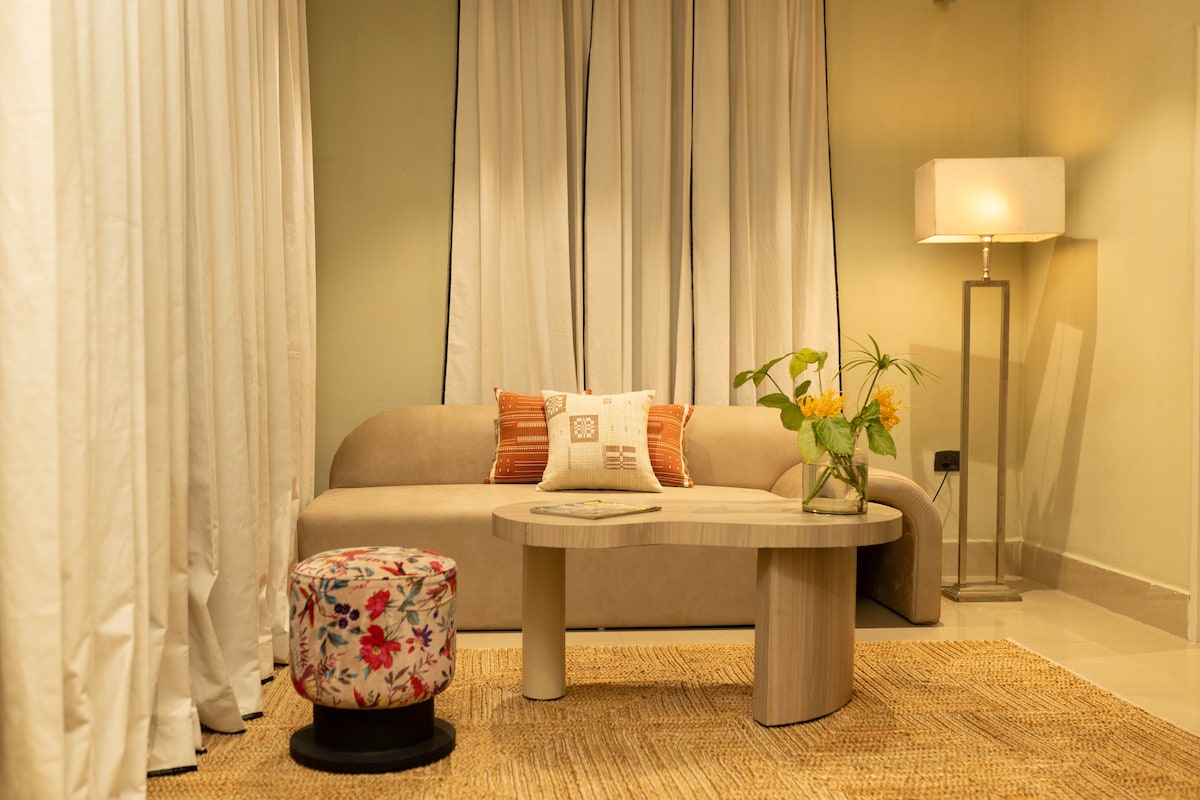 A cozy living area is showcased with a soft beige sofa adorned with decorative pillows. A round coffee table is positioned in front, accompanied by a floral-patterned stool. Sheer curtains frame the window, while a floor lamp provides gentle lighting.