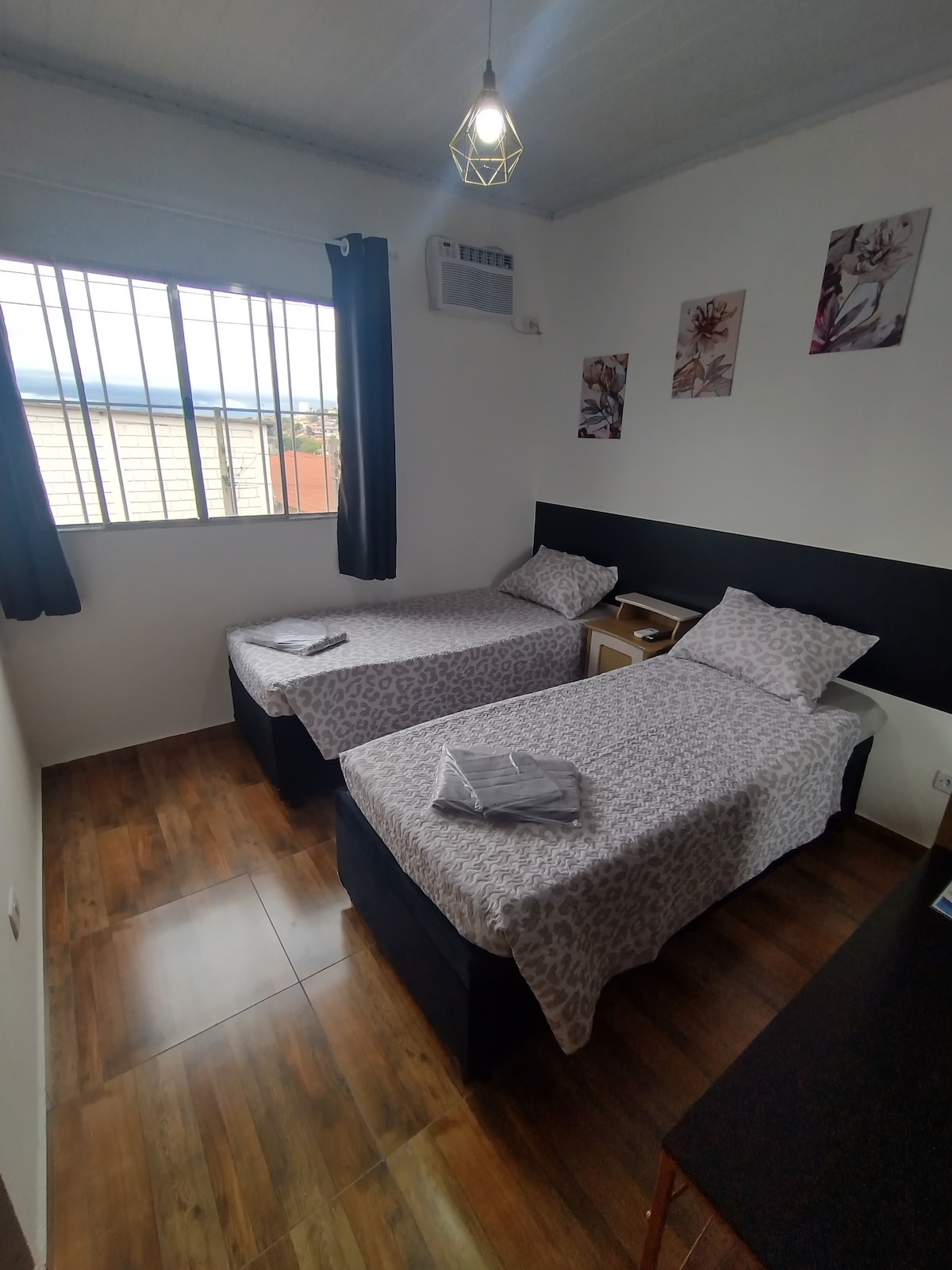 The sleeping area contains two twin beds with patterned bedding, positioned against a dark accent wall. A small nightstand is placed between them, and artwork decorates the walls. A window with black curtains allows natural light to enter, complemented by a ceiling light fixture.